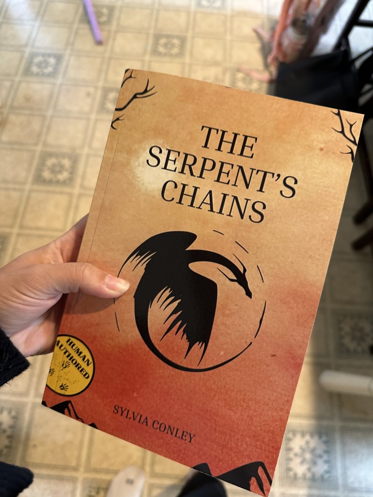 A hand holding The Serpent's Chains book - cover of a watercolor sunset background with a circular dragon graphic in the center.